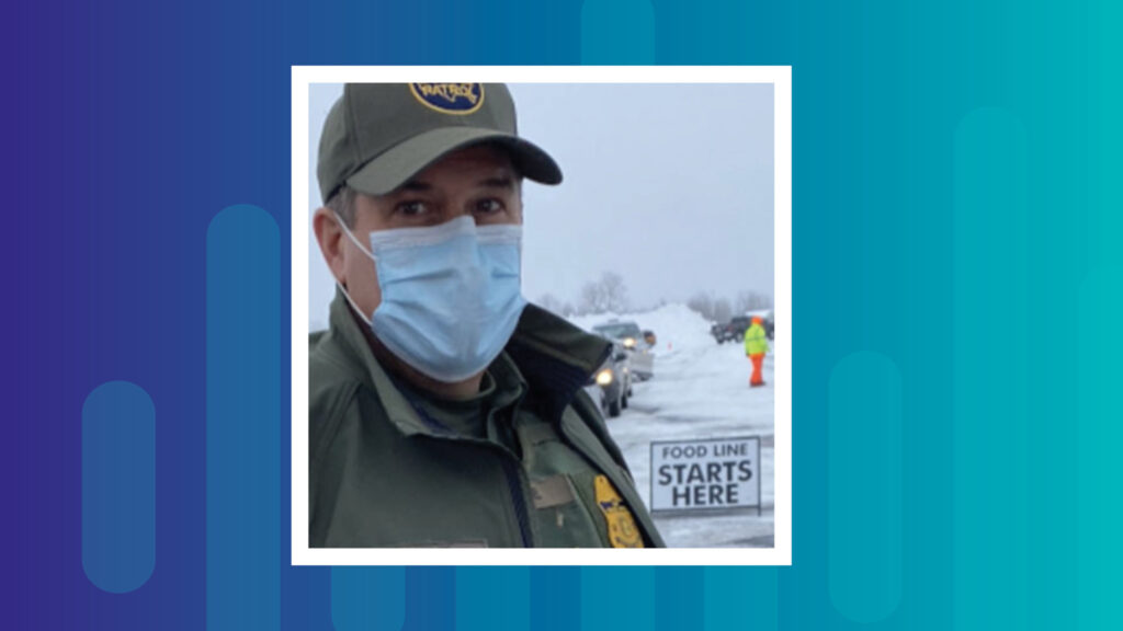 Photo of a US Border Patrol agent in Maine. He is wearing a mask, to protect from spread of COVID. There is a sign behind him that says "Food line starts here," and cars are lined up waiting. There is a fair amount of snow on the ground.