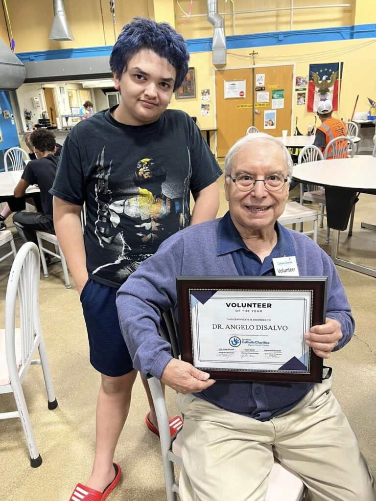 Dr. Angelo Disalvo is pictured, seated and wearing a blue sweater and khaki pants, holding an award certificate for his volunteer work as a mentor. Pictured next to him is his mentee, a boy wearing navy shorts and a black superhero t-shirt.