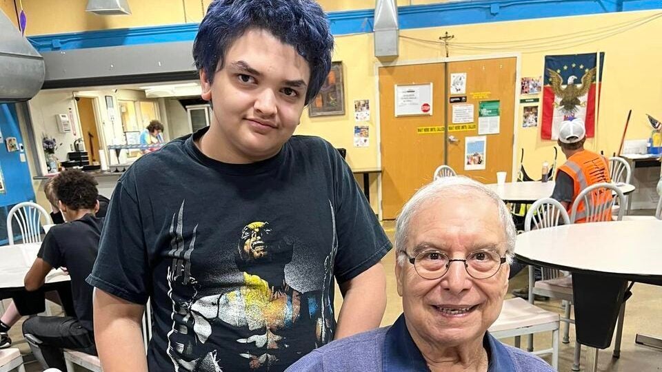 Dr. Angelo Disalvo is pictured, seated and wearing a blue sweater and khaki pants, holding an award certificate for his volunteer work as a mentor. Pictured next to him is his mentee, a boy wearing navy shorts and a black superhero t-shirt.