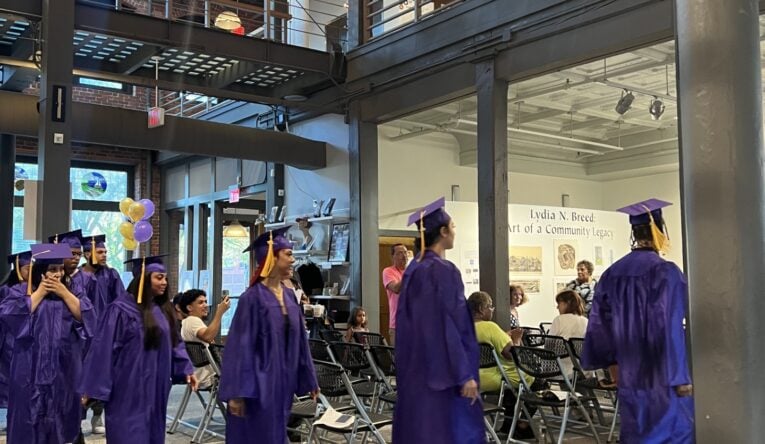 Young graduates in purple robes and caps with yellow tassels file into their high school equivalency graduation ceremony