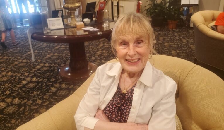 An older woman sits in a comfy chair, arms crossed, smiling at the camera. Her blonde hair is pulled back and she is wearing gold hoop earrings.