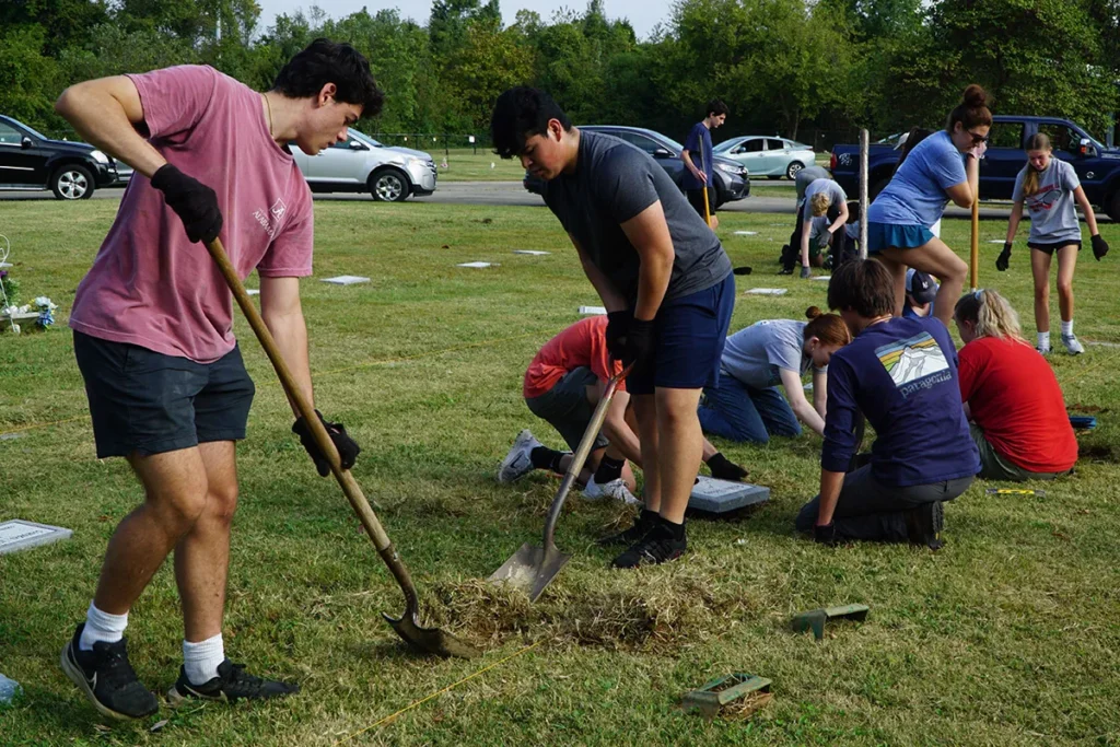 Two boys from a Louisville Catholic school, dressed in shorts and t-shirts, use shovels to dig holes for burial stones for indigent burials.