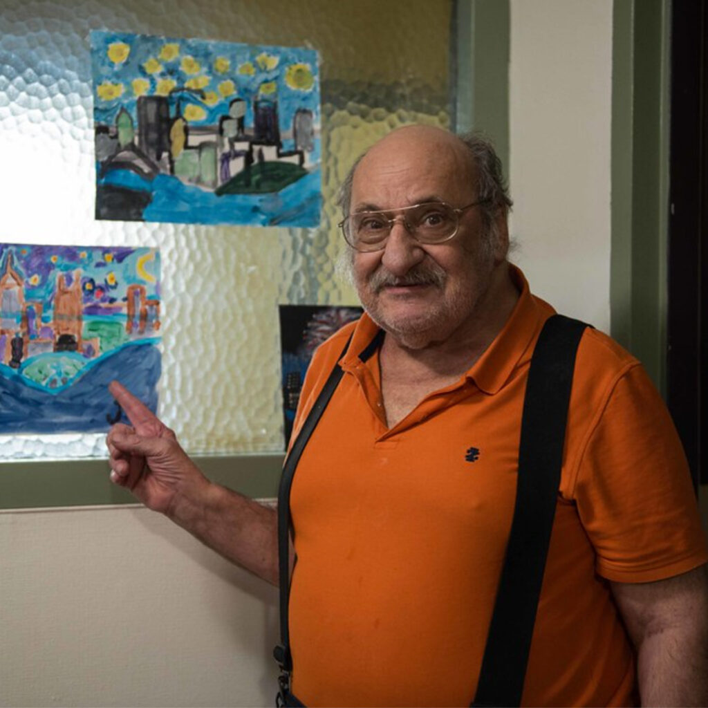 A smiling older man, wearing an orange polo shirt and suspenders, points to a painting on the wall.