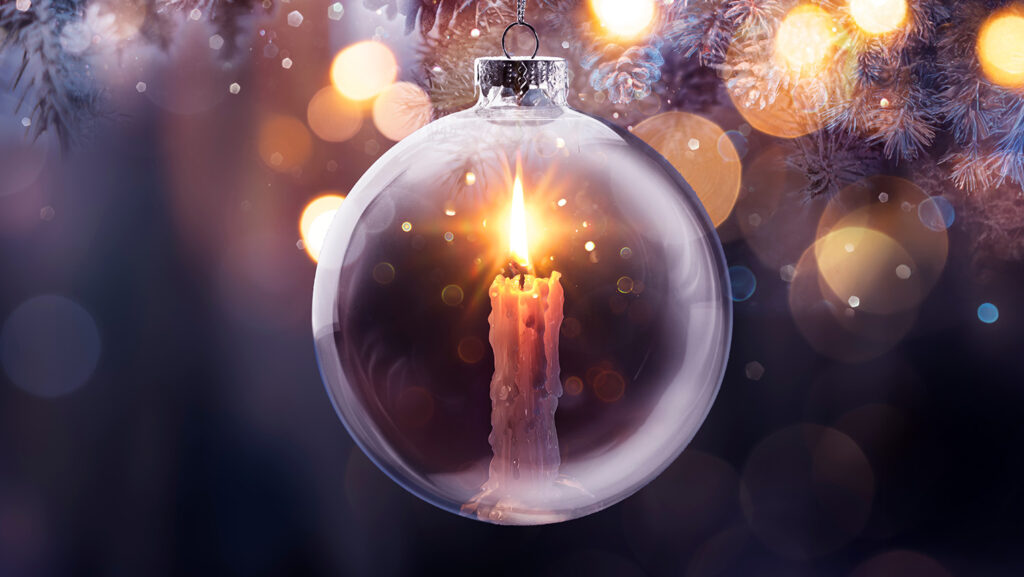 A single candle glows inside a glass ornament with soft lights glowing in the background