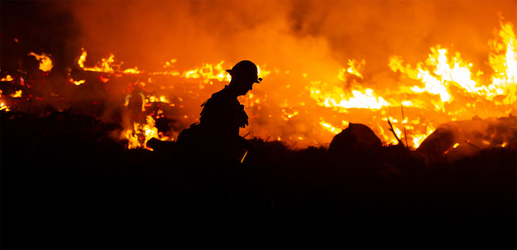 A fireman works with blazing wildfires in the background.